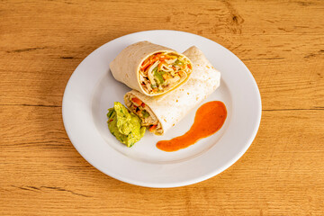 Burrito with wheat tortilla with chicken and guacamole on white plate and wooden table