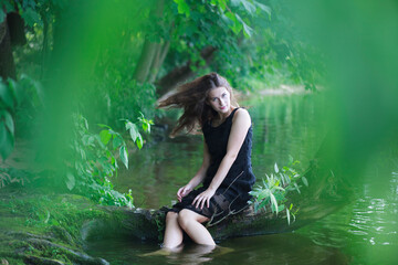 A beautiful young cheerful girl with long flowing hair in a black long dress laughs and sits on a curved tree trunk on the river bank in summer.