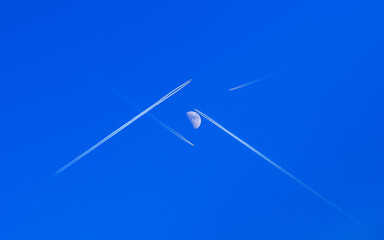 Planes fly, crossing their paths to each other directly under the moon, leaving traces behind on a clear blue sky- the true scene