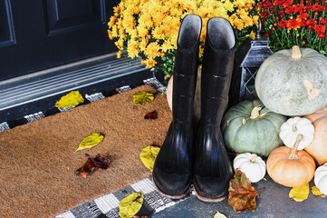 Black rain boots sitting on door mat of front porch that has been decorated for autumn with heirloom white, orange and grey pumpkins and mums. Selective focus on wellies.