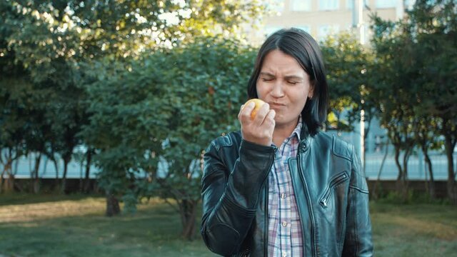 A girl stands in a city park and eats an apple. She takes a bite and realizes that the apple is not tasty. The girl winces and spits a piece of apple into her hand and then tosses it away.