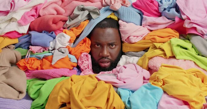 Annoyed dark skinned bearded man shouts loudly overwhelmed by domestic work covered with unfolded clothes squints face fed up of daily chores collects laundry for washing. Housekeeping concept