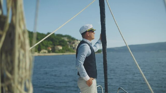 Sailor with hat standing on bow of sailboat during cruise along coast. Summer vacation at sea, sailing and travelling concept.