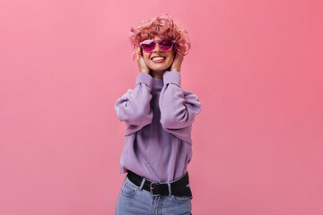 Charming young woman in purple sweater and jeans smiles sincerely. Pink-haired girl in fuchsia sunglasses poses on isolated background .