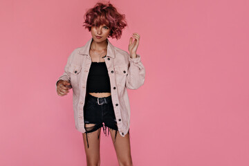 Pretty curly girl in oversized light denim jacket, black shorts and cropped dark top moves on isolated. Portrait of short-haired woman looking into camera.