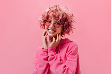 Cheerful happy woman in pink sunglasses poses on isolated background. Portrait of joyful girl in hoodie on pink backdrop.