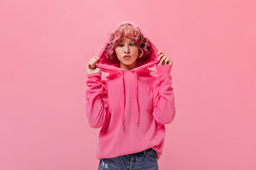 Young good-humored woman in pink hoodie makes funny face. Curly cheerful girl in denim shorts poses on isolated background.