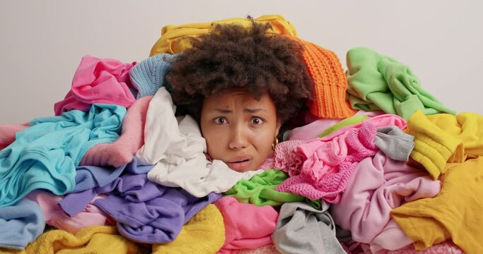 Upset displeased curly haired Afro American woman drowned in unfolded laundry poses around nessy clothes feels tired of housework isolated over white background has worried discontent expression