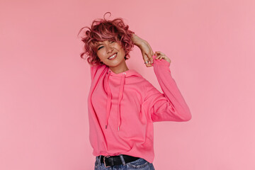 Charming woman in pink hoodie smiling on isolated. Curly cool girl in jeans poses and stretches on pink background.
