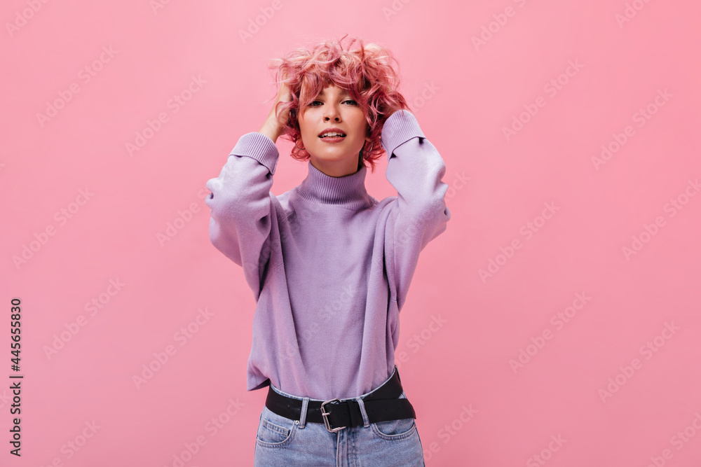 Wall mural Attractive lady in purple cashmere sweater touches curly pink hair. Young happy woman poses on isolated background.