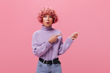 Grey-eyed woman looks surprised on isolated background. Pink-haired girl in purple sweater and jeans points to place for text on pink backdrop .