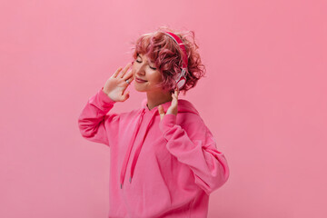 Happy young curly girl in pink hoodie dance and listens to music in headphones on isolated....