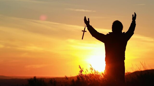 Silhouette front view of a middle-aged man holding a cross and standing on the grass with arms open to pray to God at sunset