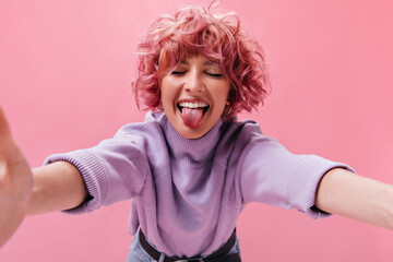 Good-humored woman in purple sweater demonstrates tongue. Pink-haired girl in cashmere shirt makes selfie on isolated .