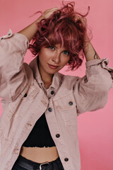 Grey-eyed charming woman ruffles pink curly hair on isolated. Tanned girl in black cropped top and denim beige jacket poses on pink background.