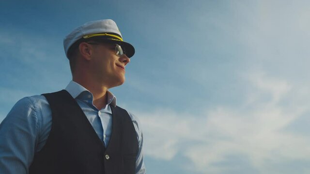 Low angle view of smiling sailor. Mid adult man with sunglasses and captain hat against sky. Summer vacation at sea, sailing and travelling concept.