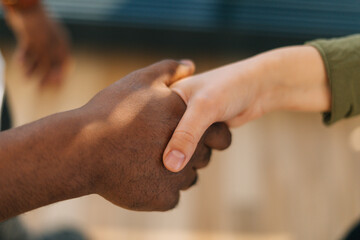 Fototapeta premium Close-up top view of handshake of Caucasian woman and black male indoors at home office, selective focus. Concept of handshaking black and white hand, interracial friendship and cooperation.