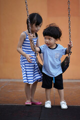 Happy Asian girl helping her little brother on swing in the playground.