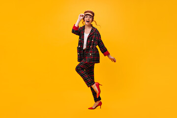 Fototapeta na wymiar Happy woman in plaid outfit moves on orange background. Cheerful girl in brown cap and eyeglasses has fan on isolated backdrop..