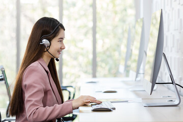 Asian young beautiful happy confident professional female operator wears pink formal suit and microphone headset sitting talking solving problem with customer while typing information with keyboard