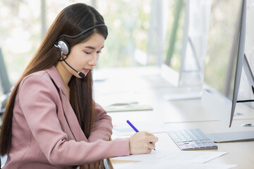 Asian young beautiful happy confident professional female operator wears pink formal suit and microphone headset sitting talking solving problem with customer while typing information with keyboard