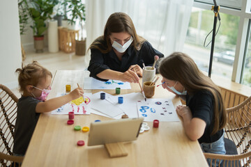 a girl art teacher sits in the center of a wooden table during the day in a classroom with stained glass windows in front of a tablet computer and teaches children to draw medical masks on everyone's