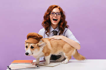 Optimistic woman in eyeglasses smiles and hugs corgi. Funny student girl in shirt plays with dog in...