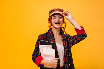 Curly woman in cap and eyeglasses smiles and holds notebooks. Lovely young girl with red hair in...