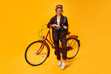 Naklejka premium Woman in plaid suit leans on bicycle. Stylish wonderful girl in cool clothes, white sneakers and brown cap posing on isolated background..