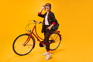 Beautiful woman in plaid outfit and cap posing with bicycle. Fashionable girl in eyeglasses and modern suit looking into camera..