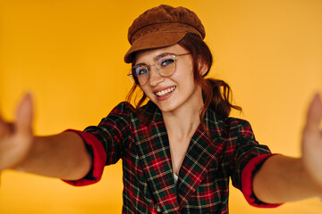Pretty woman in trendy headwear takes selfie on orange background. Charming girl in brown cap and eyeglasses makes photo and smiling..