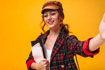 Cheerful lady in eyeglasses taking selfie and holding notebooks. Stylish curly woman in brown cap and plaid suit makes photo..