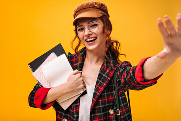 Pretty girl in cap and plaid jacket smiling and holding notebooks on orange background. Beautiful woman in eyeglasses and bright suit posing..