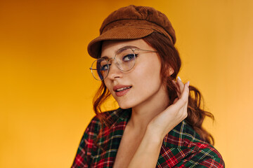 Naklejka premium Ginger lady in stylish eyeglasses poses on orange background. Cool woman with red wavy hair in brown trendu cap looking into camera..