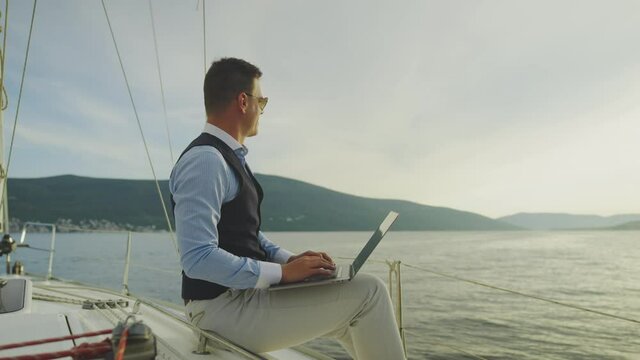 Businessman working outdoors, sitting at sailboat and typing on laptop. Summer vacation at sea, sailing and travelling concept.