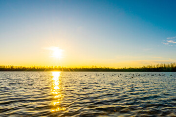 The setting sun shines on the calm lake with golden sunlight reflected from the rippling surface. A beautiful sunset