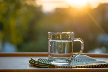 Clean drinking water in a glass cup with a handle stands on a napkin on a wooden table against the background of green nature.Sunset. The topic is thirst quenching, ecology. Selective focus