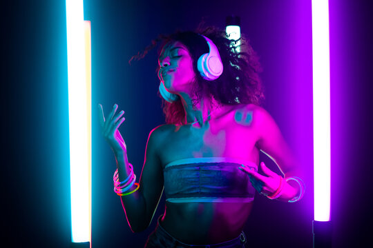 Portrait of party girl dancing to music with headphones on glowing multi-colored lamps in studio. Disco woman with curly hairstyle, modern outfit. Amazing style.