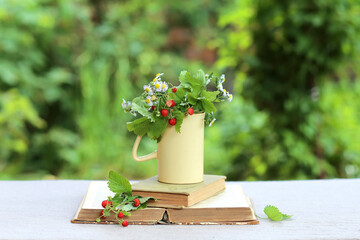 A strawberry bush in a yellow cup and a bouquet of daisies on a stack of books on a table in the garden. Summer leisure and relaxation. Vertical photo background