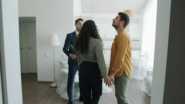 Happy real estate agent in suit is showing new house to couple of buyers talking gesturing making property presentation. Business and people concept.