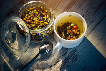 Dry mix of tea leaves, herbs, petals, berries and nuts in glass bowl. Natural herbal tea in white cup on gray wooden background in the evening light, shadow on the table.