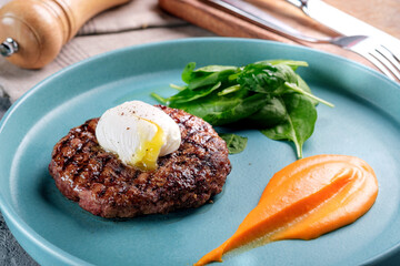 Minced beef steak with poached egg. Steak dish on a blue plate with spinach and pumpkin