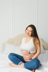 Portrait of nice adorable lovely sweet tender beautiful cheerful joyful pregnant curly-haired mom in casual wear sitting on divan, holding belly, white light interior room