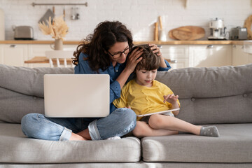 Loving mother help preschool kid with e-learning on digital tablet work on laptop computer from home. Caring young mom embrace little son playing games on sofa. Parent and child on lockdown concept