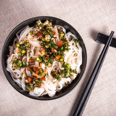 Rice noodles with herbs, vegetables and soy sauce in a bowl with chopsticks on a linen napkin, close-up, top view