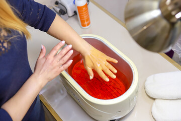 Paraffin theoraphy. Female hand and orange paraffin wax in bowl. Manicure and skincare. Woman girl in beauty spa salon.