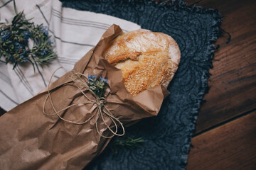 Sourdough bread sprinkled with sesame seeds wrapped in paper bag decorated with rosemary flowers. Freshly baked bread placed on dark wooden surface.