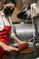 Beautiful young female seller with protective face mask working in a confectionery shop or bakery. She is making delicious sweets. Coronavirus and small business concept.