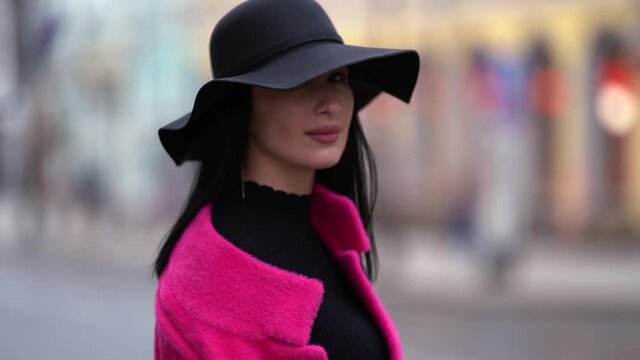 portrait of a brunette in a black hat and a bright pink coat against the background of a blurred city street. the camera is moving