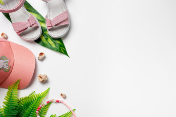 Top view with summer accessories. Girl's accessories, pink cap and sandals with green tropical leaves and shells on the white isolated background . 